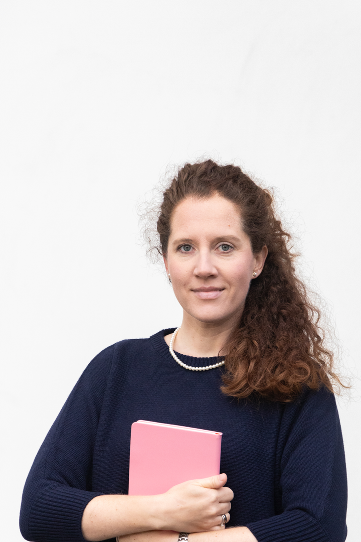Nicole-Kristin vor einer weißen Wand mit rosa Buch im Arm.
